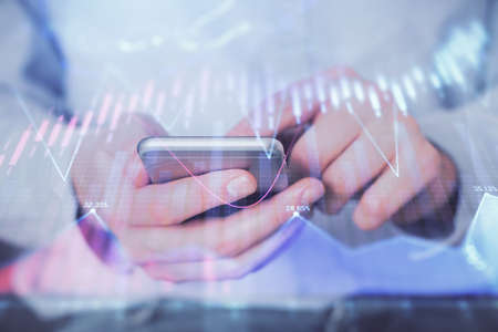 Double exposure of man's hands holding and using a digital device and forex graph drawing. Financial...