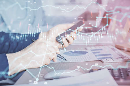 Double exposure of man's hands holding and using a digital device and forex graph drawing. Financial...
