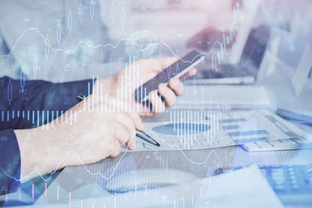 Double exposure of man's hands holding and using a digital device and forex graph drawing. Financial...