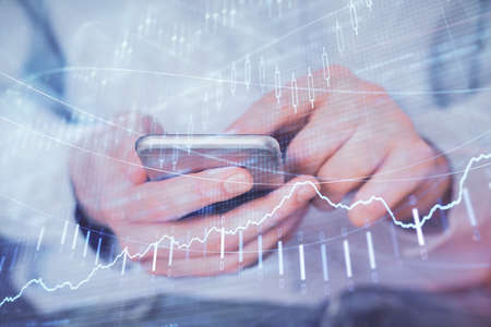 Double exposure of man's hands holding and using a digital device and forex graph drawing. Financial...