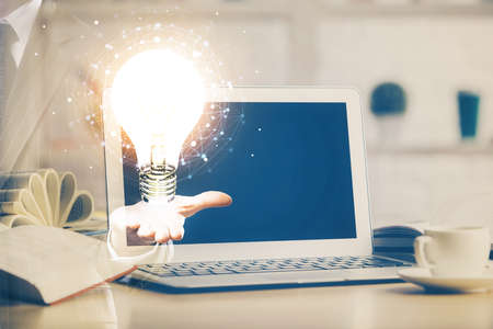 Computer on desktop in office with bulb icon hologram. Double exposure. Concept of idea.