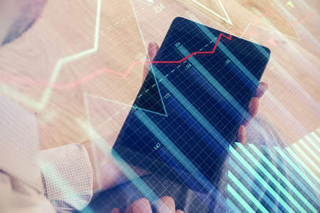 Double exposure of man's hands holding and using a digital device and forex graph drawing. Financial...
