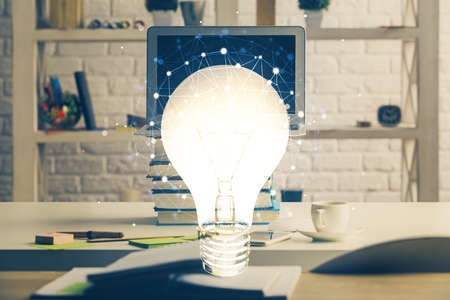 Computer on desktop in office with bulb icon hologram. Double exposure. Concept of idea.