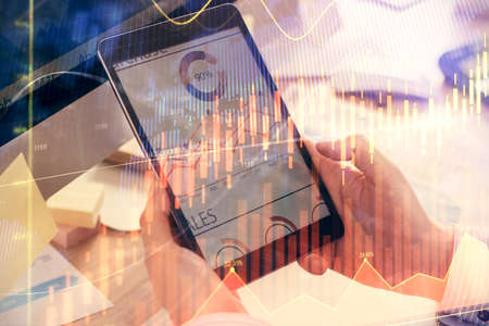 Double exposure of mans hands holding and using a phone and financial graph drawing. Analysis...