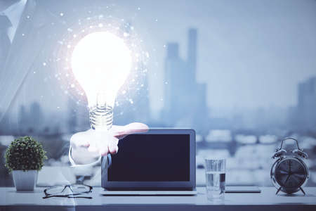 Computer on desktop in office with bulb icon hologram. Double exposure. Concept of idea.