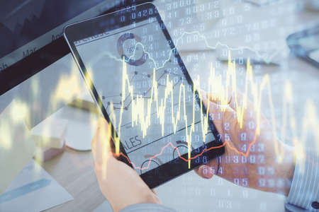 Double exposure of mans hands holding and using a phone and financial graph drawing. Analysis...