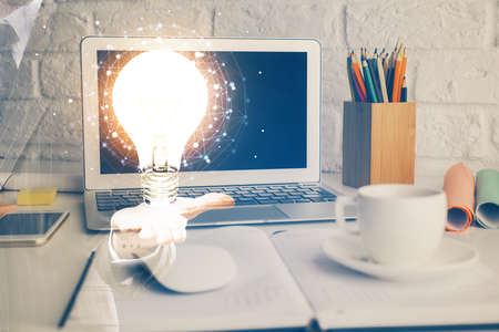 Computer on desktop in office with bulb icon hologram. Double exposure. Concept of idea.
