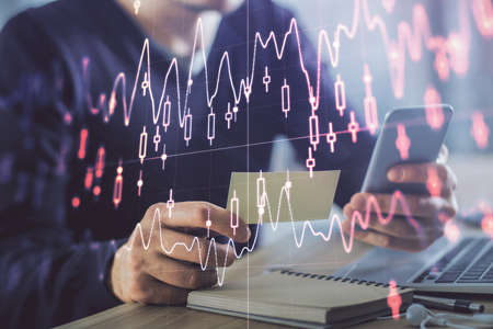 Double exposure of mans hands holding and using a phone and financial graph drawing. Analysis...