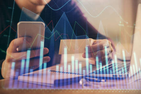 Double exposure of man's hands holding and using a phone and financial graph drawing. Analysis...