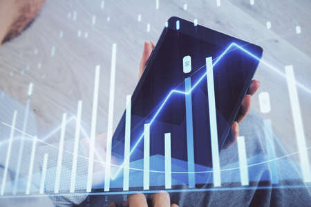 Double exposure of man's hands holding and using a digital device and forex graph drawing. Financial...