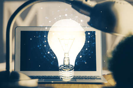 Computer on desktop in office with bulb icon hologram. Double exposure. Concept of idea.