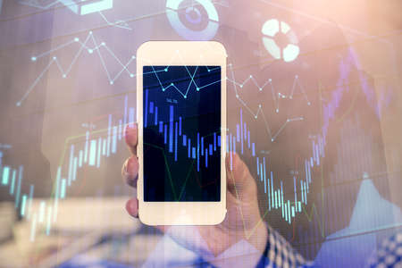 Double exposure of mans hands holding and using a digital device and forex graph drawing. Financial...