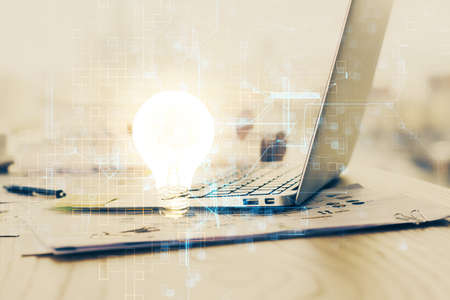 Computer on desktop in office with bulb icon hologram. Double exposure. Concept of idea.