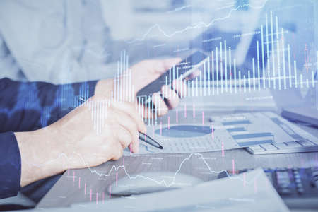 Double exposure of mans hands holding and using a digital device and forex graph drawing. Financial...