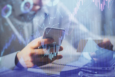 Double exposure of mans hands holding and using a phone and financial graph drawing. Analysis...