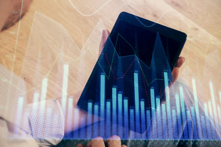 Double exposure of mans hands holding and using a digital device and forex graph drawing. Financial...