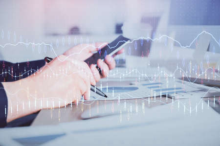 Double exposure of mans hands holding and using a phone and financial graph drawing. Analysis...