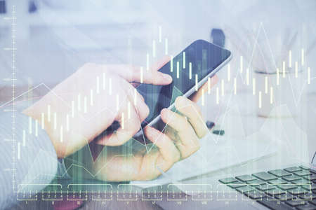 Double exposure of mans hands holding and using a digital device and forex graph drawing. Financial...