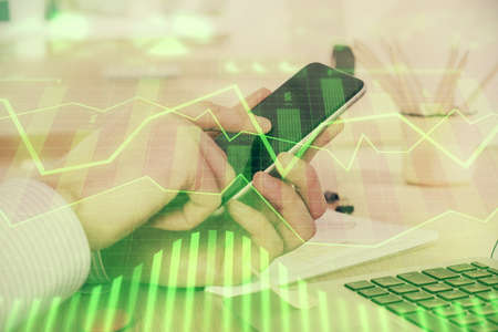 Double exposure of mans hands holding and using a digital device and forex graph drawing. Financial...