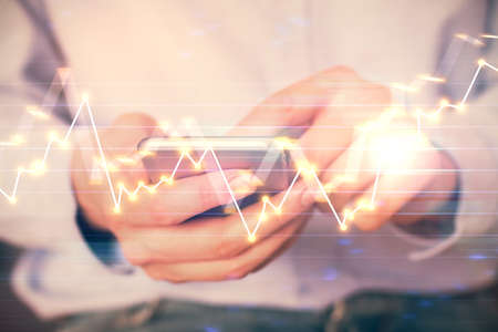 Double exposure of mans hands holding and using a digital device and forex graph drawing. Financial...