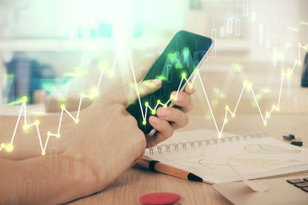 Double exposure of mans hands holding and using a digital device and forex graph drawing. Financial...