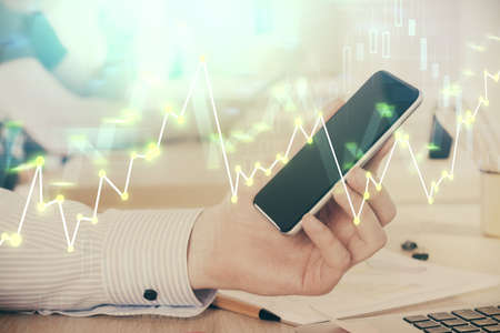 Double exposure of man's hands holding and using a digital device and forex graph drawing. Financial...