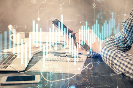 Double exposure of man's hands holding and using a digital device and forex graph drawing. Financial...