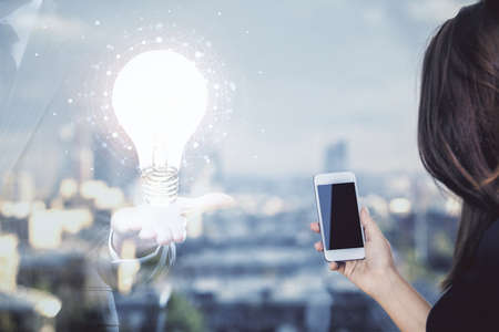 Double exposure of light bulb hologram and woman holding and using a mobile device. Idea concept.の写真素材
