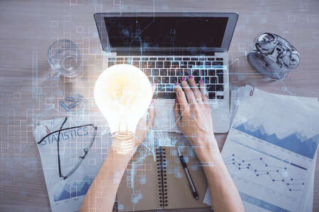 Double exposure of woman hands working on computer and light bulb hologram drawing. Top View. Idea...