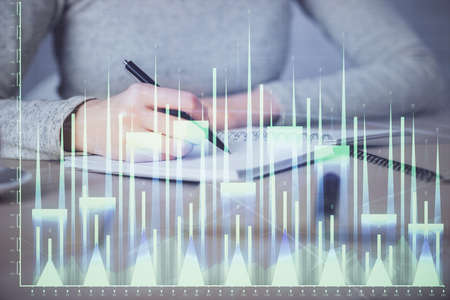 Hands writing information about stock market in notepad. Forex chart holograms in front. Concept of...