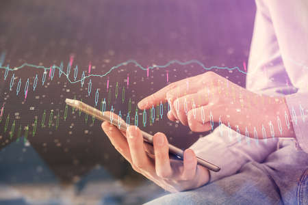 Double exposure of man's hands holding and using a digital device and forex graph drawing. Financial...