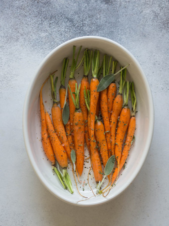 Fresh peeled orange carrots with fresh sage and dry thyme in a white ceramic baking sheet on gray background. Top viewの写真素材
