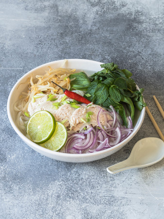 Traditional Vietnamese soup- pho ga in white bowl with chicken and rice noodles, mint and cilantro, green and red onion, chili, bean sprouts and lime on grey background.の写真素材