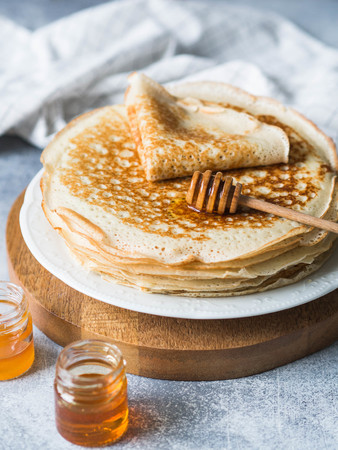 Stack Russian homemade yeast pancakes on white plate, wood honey spoon and honey in jars on table.