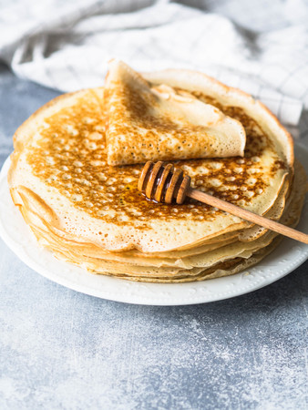 Stack Russian homemade yeast pancakes on white plate, wood honey spoon on table.の写真素材