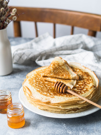 Stack Russian homemade yeast pancakes on white plate, wood honey spoon and honey in jars on grey table.の写真素材
