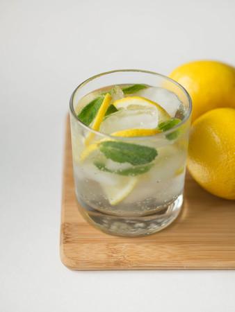Summer citrus lemonade with ice in glass on wood board and two lemons on whiteの写真素材