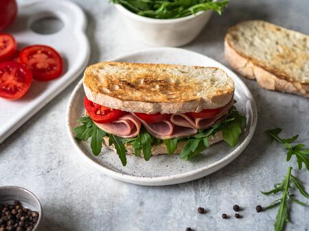 Ham, tomato and arugula sandwich on toasted bread slices on a white plate and other ingredients on a gray backgroundの写真素材