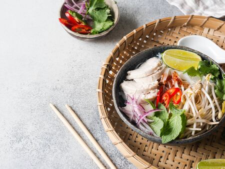 Traditional Vietnamese soup- pho ga in bowl with chicken and rice noodles, mint and cilantro, red onion, chili, bean sprouts and lime on grey background. Asian food.の写真素材