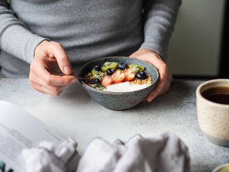 Uncertain girl in grey dress at the table with breakfast. Bowl with yogurt, granola and berries and...