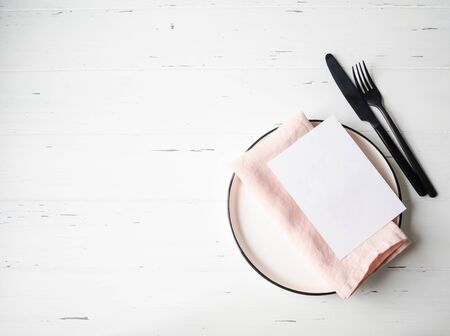 Rustic table setting with plate, pink napkin, paper card and appliances on white wood table.の写真素材