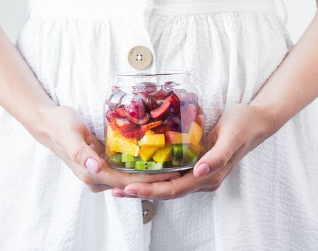 Girl hold glass jar with berries and fruits. Fruit salad with kiwi, peach, strawberry, cherries and plum in glass jar on white napkin. Summer seasonal tasty vegetarian salad for snack or lunchの写真素材