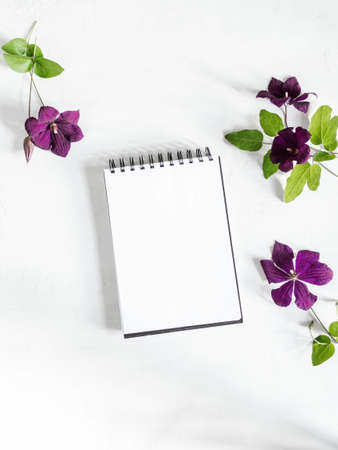 Flat lay composition of purple clematis flowers and leaves and notepad with a blank sheet layout on...