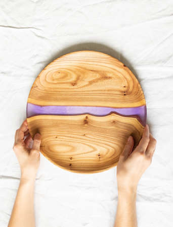 Female hands holding round wooden craft tray with a lilac resin insert on a white textile background. Top view. Copy spaceの写真素材