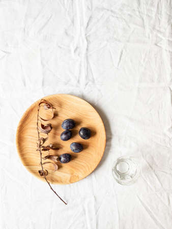 Wooden homemade round plate with plums and dry branch with leaves on a white cloth background. Top view. Copy spaceの写真素材