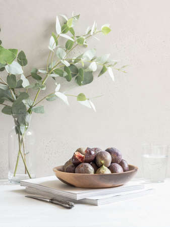 Minimalistic still life with a wooden plate full of ripe figs and a glass of water against a light...