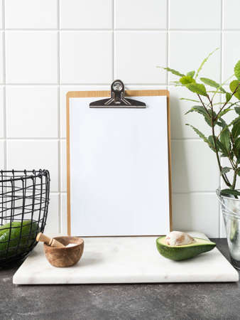 Stylish white kitchen background with basket with avocado, laurel plant in pot and clipboard with blank sheet of paper for text, copy space, front viewの写真素材