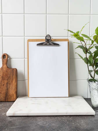 Stylish white kitchen background with cutting board, laurel plant in pot and clipboard with blank sheet of paper for text, copy space, front viewの写真素材