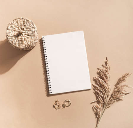 White blank notepad sheet with spiral, sprigs of reeds, gold earrings and wicker box on pastel beige background. top view. Copy space. Flat layの写真素材