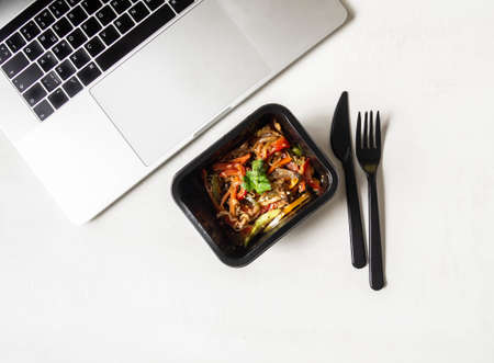 Black plastic container with vegetables stir fry and black appliances and laptop on white...